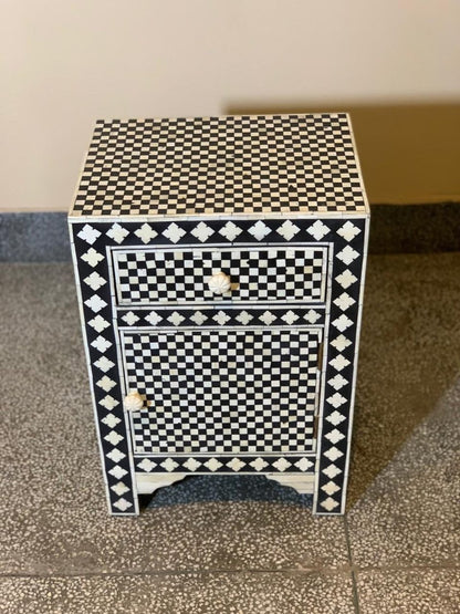 Handcrafted bone inlay nightstand in black and white checker pattern with diamond border, one drawer with carved knob, lower cabinet door, angled view showing top and front panels