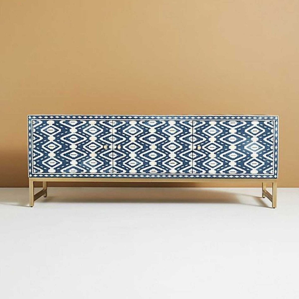 Front angle of a handmade indigo and cream bone inlay entertainment unit, showing the geometric ikat pattern across the cabinet front, three storage sections with solid brass handles, and the long rectangular top supported by a slim brass base.