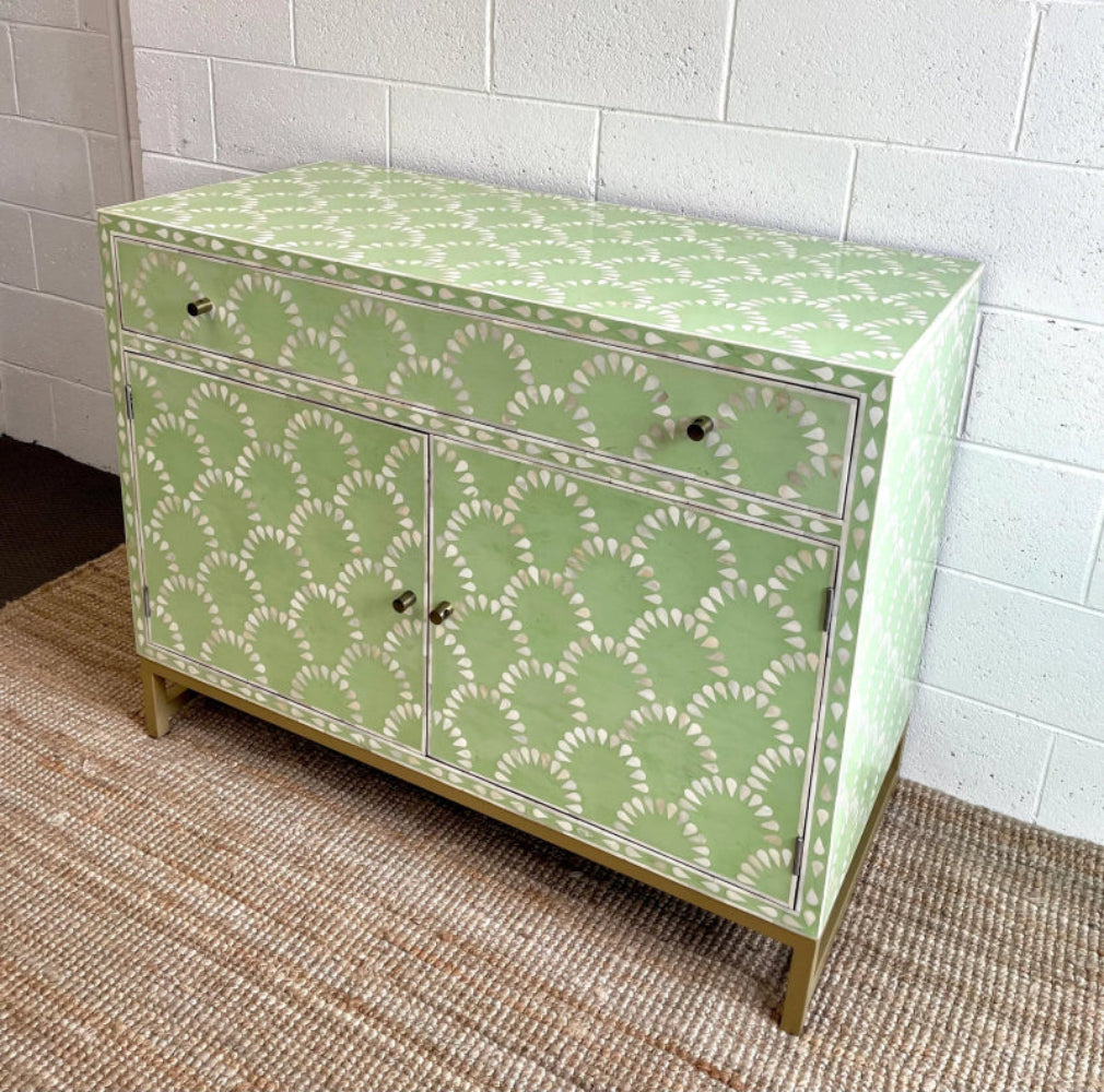 Angled view of a sage green bone inlay scalloped cabinet showing top surface, drawer, two doors with brass knobs, and gold metal base on a jute rug