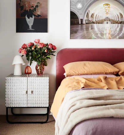 Grey bone inlay two-door bedside cabinet on black sled base styled in a warm bedroom with pink roses, yellow bedding, and wall art