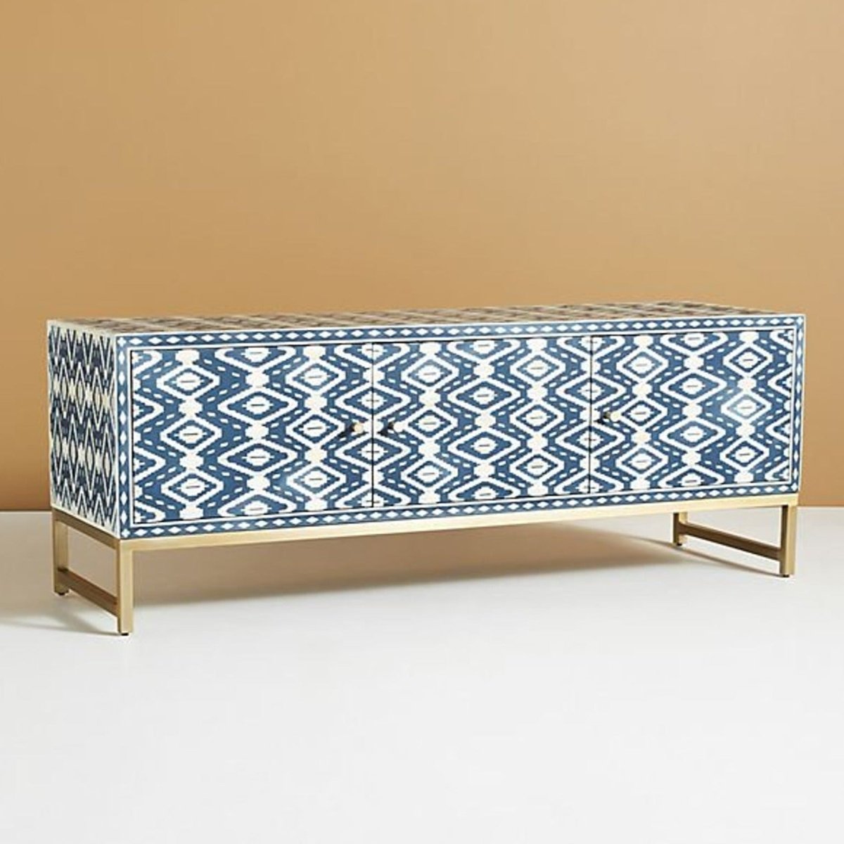 Full front view of a handmade indigo and cream bone inlay media console, showing the repeating ikat pattern across three storage doors, solid brass handles, a straight rectangular top, and the raised brass base against a plain background.