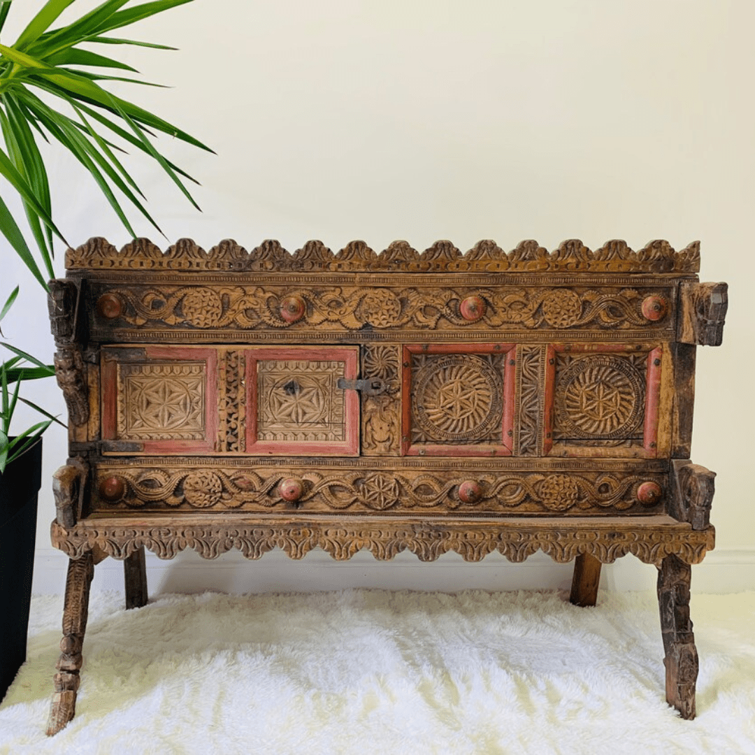 Natural wood hope chest, front view showing red-accented carved panels, scalloped borders and legs on a white rug.