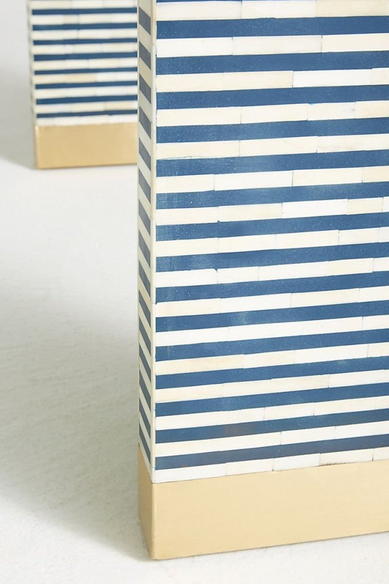 Side profile close-up of the handmade navy and white bone inlay table, showing horizontal stripes across the side panel, pale inlay seams, a rounded edge, and the gold-tone block foot supporting the curved waterfall construction.