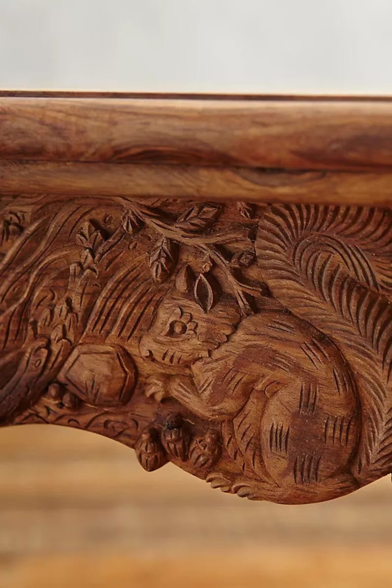Ornate solid wood kitchen table in a warm finish, edge detail view showing animal carvings across the apron.