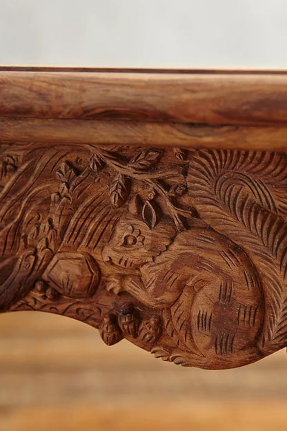 Ornate solid wood kitchen table in a warm finish, edge detail view showing animal carvings across the apron.