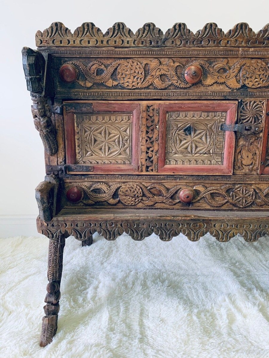 Red-accented carved hope chest in natural wood, front view cropped to the left panels, latch and carved leg.