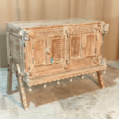 Rustic two-door wood server in natural tones, front view at an angle showing the left side, carved trim and legs.