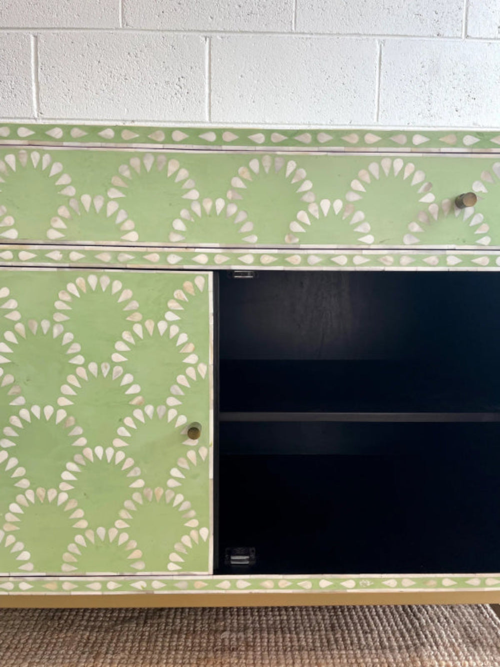 Sage green scalloped bone inlay cabinet with one door open showing dark interior shelf, one closed door with brass knob, gold metal base on jute rug