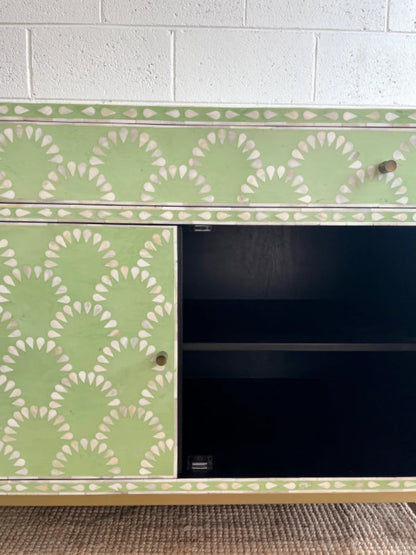 Sage green scalloped bone inlay cabinet with one door open showing dark interior shelf, one closed door with brass knob, gold metal base on jute rug