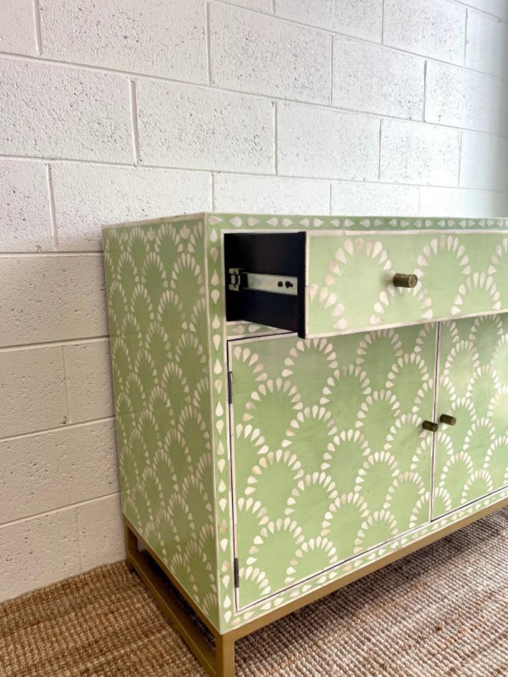Sage green scalloped bone inlay cabinet with drawer open showing dark interior and hardware, two closed doors with brass knobs, gold metal base on jute rug