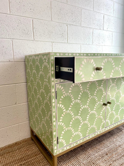 Sage green scalloped bone inlay cabinet with drawer open showing dark interior and hardware, two closed doors with brass knobs, gold metal base on jute rug