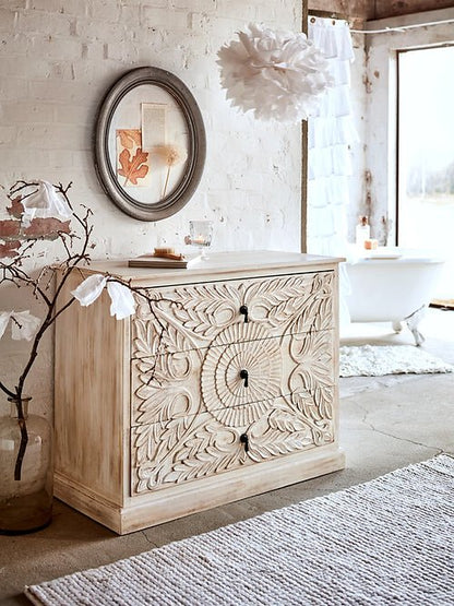 A 3-drawer solid wood dresser with intricate hand-carved floral and leaf patterns in a natural wood finish, featuring black teardrop pulls.