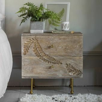 Two-drawer nightstand in a light, weathered wood finish with a continuous hand-carved fern leaf pattern across the drawers, standing on sleek brass legs with ball feet.
