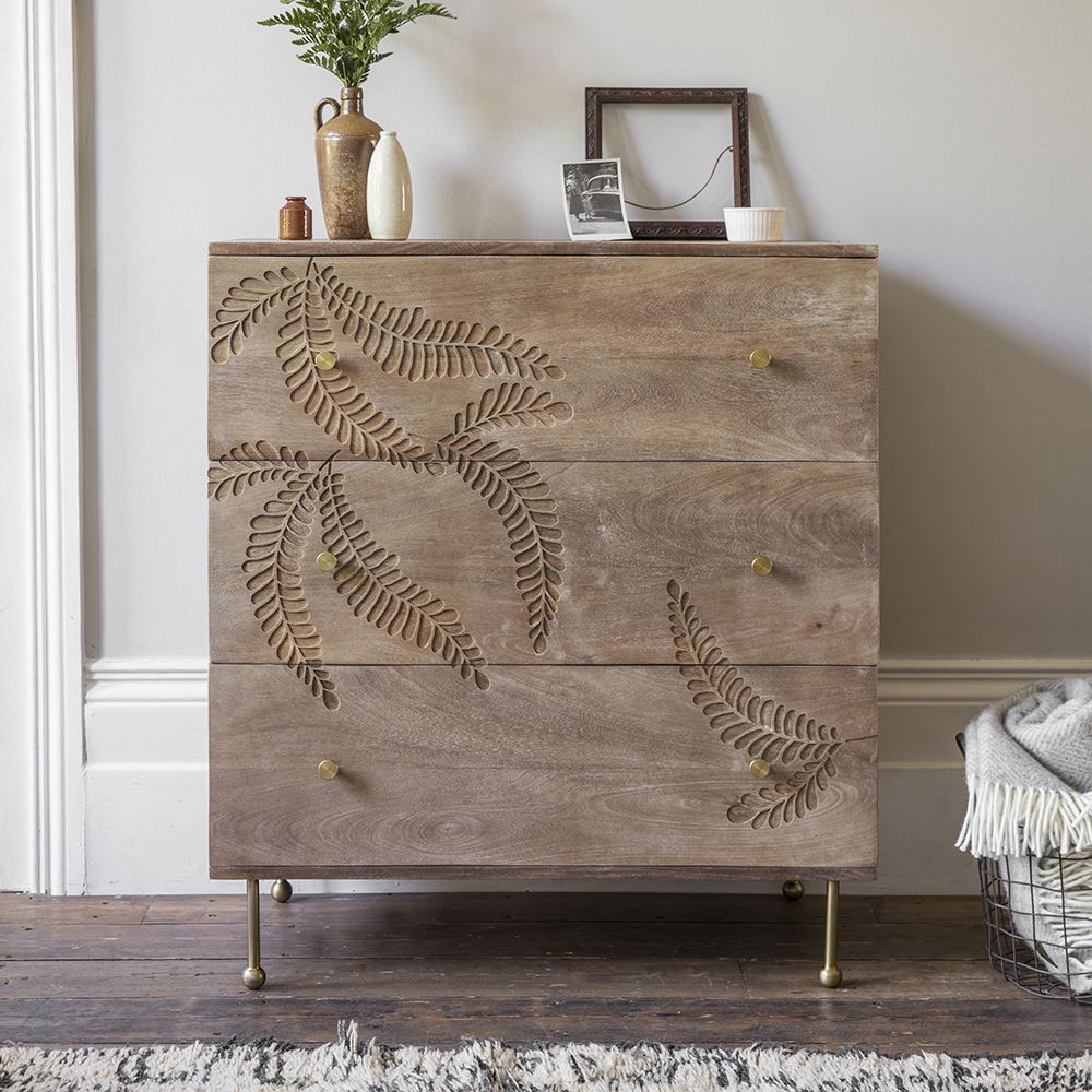 A 3-drawer solid wood dresser with an asymmetrical hand-carved fern leaf pattern on the front, featuring gold-toned metal legs and hardware.
