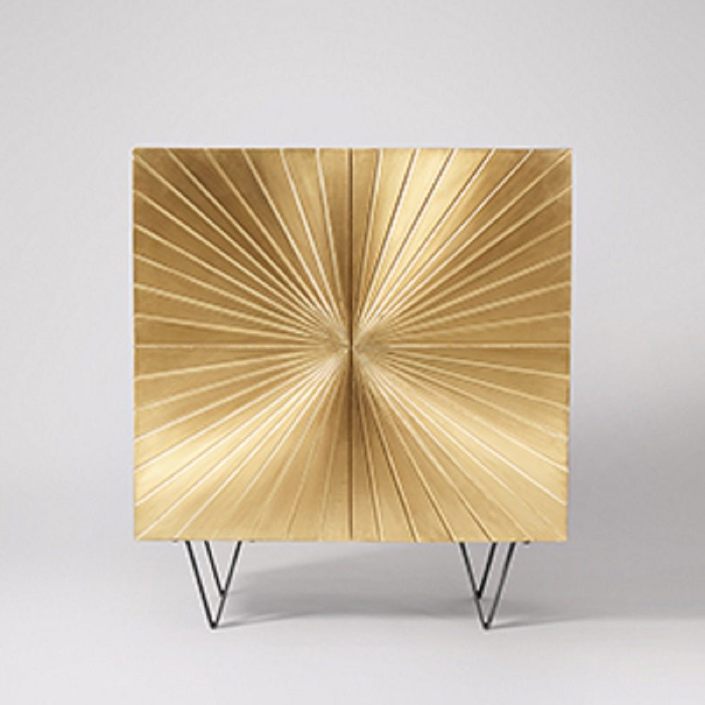 Ziggy two-door square bar cabinet wrapped in a brushed brass gold finish with a large central sculpted sunburst pattern on black metal hairpin legs.