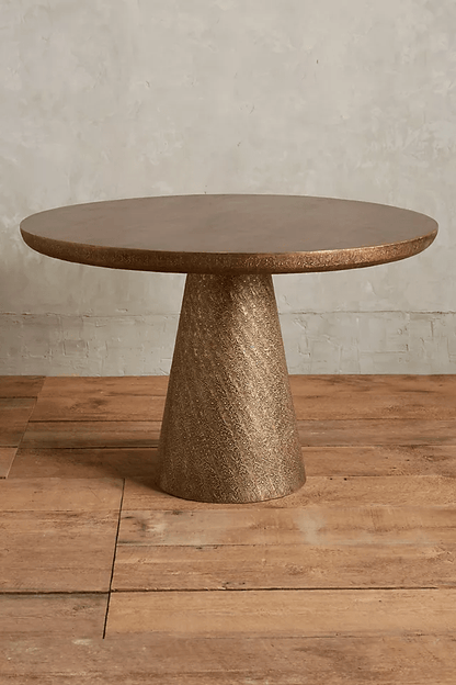 Round dining table featuring a smooth brown wood top with a bronze textured edge on a large conical hand-embossed metal base resting on wood floors.