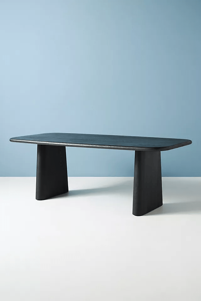 Modern Kalle dining table in matte black solid wood featuring thick tapered pedestal legs and rounded rectangular top against a blue and white background.