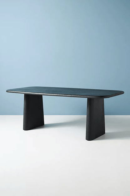 Modern Kalle dining table in matte black solid wood featuring thick tapered pedestal legs and rounded rectangular top against a blue and white background.