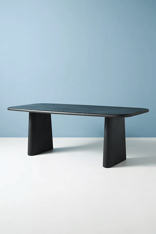 Modern Kalle dining table in matte black solid wood featuring thick tapered pedestal legs and rounded rectangular top against a blue and white background.