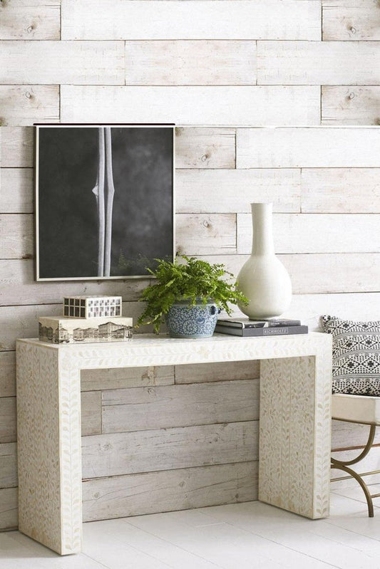 White floral bone inlay waterfall console table styled with a white vase, books, and greenery on a rustic white plank wall.